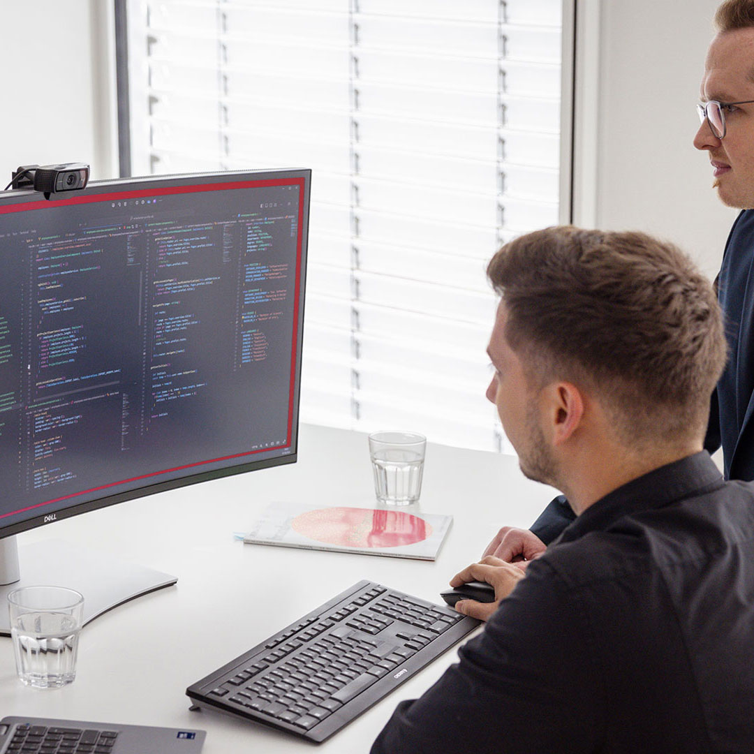 Ein Programmierer und sein Kollege der DOS beraten sich zu einer individuellen IT-Lösung vor dem Monitor